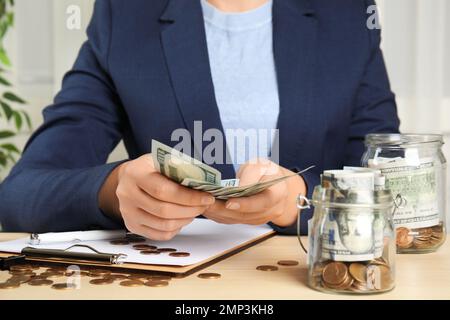 Eine Frau, die Geld in der Nähe eines Glasbehälters zählt, mit Dollarscheinen und Münzen auf einem Holztisch, Nahaufnahme Stockfoto