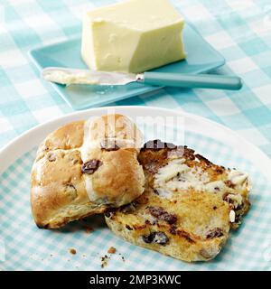 Heiße Kreuzbrötchen gebuttert in Scheiben geröstete ostern traditionelle Gingham Tischtuch Küchenmesser Butterschmelze köstlicher saisonaler Weizenboden gebackener Kuchen Stockfoto