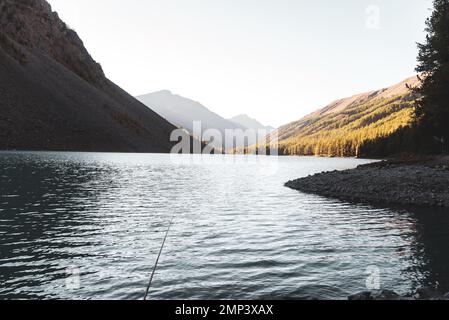 Eine Angel liegt im Schatten auf den Felsen am Ufer des alpinen Sees Shavlinskoe am Abend mit Sonnenlicht in den Bergen am Horizont. Stockfoto