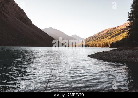 Eine Angel liegt im Schatten auf den Felsen am Ufer des alpinen Sees Shavlinskoe mit Sonnenlicht in den Bergen am Horizont in Altai. Stockfoto