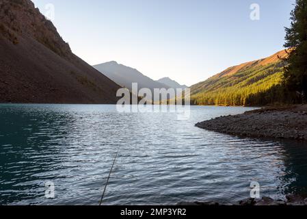 Eine Angelrute liegt im Schatten auf den Steinen am Ufer des alpinen Sees Shavlinskoye mit Sonnenlicht am Abend bei Sonnenuntergang in den Bergen weiter Stockfoto