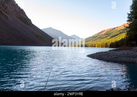 Eine Angel liegt im Schatten auf den Felsen am Ufer des alpinen Sees Shavlinskoe mit Sonnenlicht in den Bergen am Horizont. Stockfoto