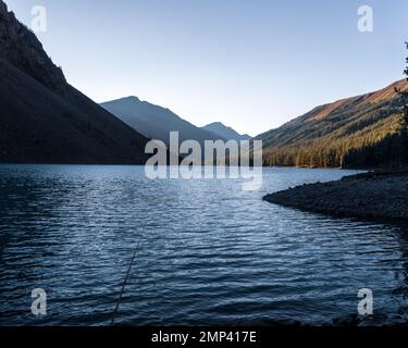 Eine Angelrute liegt im Schatten auf den Felsen am Ufer des alpinen Sees Shavlinskoye mit Sonnenlicht bei Sonnenuntergang in den Bergen am Horizont. Stockfoto