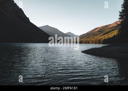 Eine Angelrute liegt im Schatten auf den Felsen am Ufer des alpinen blauen Sees Shavlinskoe mit Sonnenlicht bei Sonnenuntergang in den Bergen am Horizont. Stockfoto