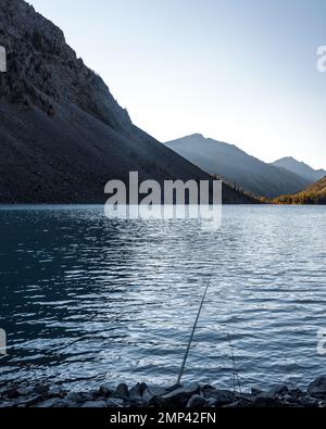 Eine Angelrute liegt im Schatten auf den Felsen am Ufer des alpinen Sees Shavlinskoye mit Sonnenlicht bei Sonnenuntergang in den Bergen am Horizont. Stockfoto