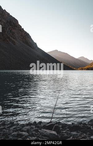 Eine Angelrute liegt im Schatten auf den Felsen am Ufer des alpinen Sees Shavlinskoye mit Sonnenlicht bei Sonnenuntergang in den Bergen am Horizont. Ver Stockfoto