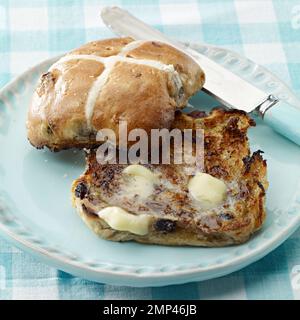 Heiße Kreuzbrötchen gebuttert in Scheiben geröstete ostern traditionelle Gingham Tischtuch Küchenmesser Butterschmelze köstlicher saisonaler Weizenboden gebackener Kuchen Stockfoto