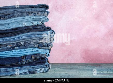 Stapel verschiedener Jeans auf blauem Holztisch vor pinkfarbenem Hintergrund. Platz für Text Stockfoto