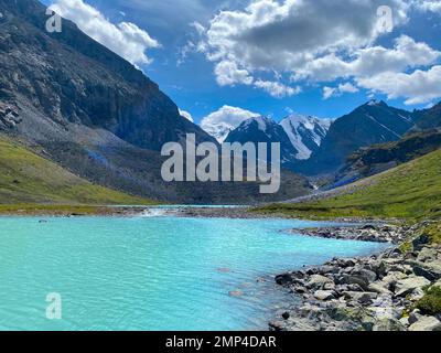 Bergsee mit türkisfarbenem Wasser Karakabak im Altai-Gebirge mit schneebedeckten Gipfeln und Gletschern unter den Wolken und grünem Gras mit Hängevorrichtung Stockfoto