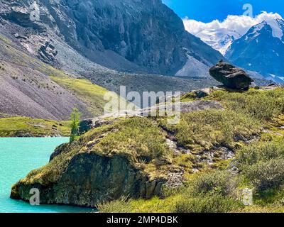 Türkisfarbenes klares Wasser des Karakabak-Sees im Altai-Gebirge mit Berggipfeln unter Wolken und grünem Gras mit hängenden Steinen. Stockfoto