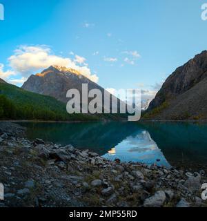 Panorama des Ufers des Shavlinskoe-Sees im Schatten mit Steinen inmitten der Berge mit einer Reflexion der Gipfel mit Gletschern und Schnee in Altai. Stockfoto