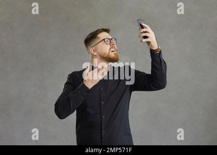 Erstaunter und verwirrter junger Mann sieht frustriert aus, wenn er den Smartphone-Bildschirm in seinen Händen sieht. Stockfoto