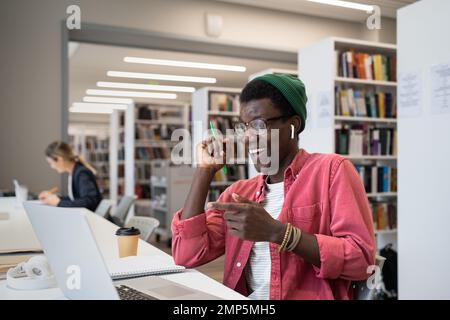 Positiver junger afroamerikanischer Lehrer mit Ohrstöpseln, der Online-Unterricht in der Bibliothek führt Stockfoto