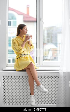 Junge Frau mit Becher auf Fensterbank drinnen Stockfoto