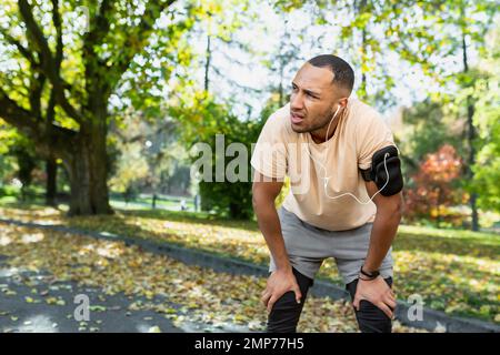 Ein junger afroamerikanischer Sportler hört Musik in weißen Kopfhörern und geht morgens im Park joggen. Er ist müde, hat angehalten, atmet schwer, steht und ruht sich aus. Stockfoto