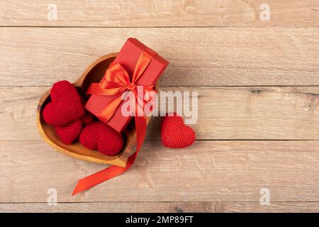 Rote gestrickte Herzen und Geschenkschachteln in einer Holzschüssel in Herzform auf Holzhintergrund. Happy Valentine's Day, Mother's Day and Birthday gr. (Alles gute zum Valentinstag, Muttertag und Geburtstag Stockfoto