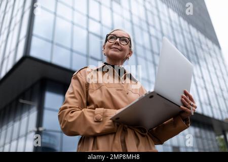 Grauhaarige, reife, europäisch aussehende Geschäftsfrau mit einem Laptop vor dem Hintergrund der Glasfassade des Büros Stockfoto