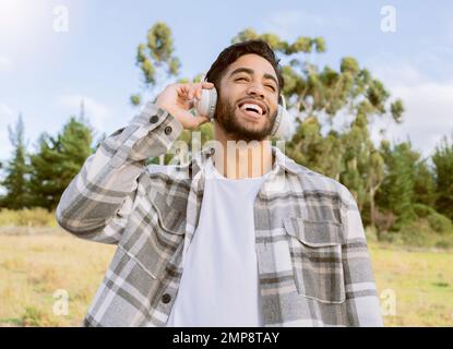 Musik, Freude und Mann entspannen Sie in einem Park, lächeln und lachen Sie beim Podcast oder Audiotrack vor dem Hintergrund der Natur. Kopfhörer, lachen und gutaussehender Kerl Stockfoto