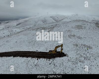 Hydraulikbagger mit Hydraulikhammer, der auf einem verschneiten Berg mit Windturbinen arbeitet. Stockfoto