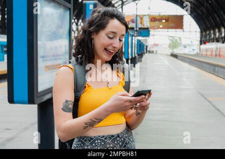 Junge lateinin argentinischer ethnischer Herkunft, die gelb gekleidet ist, steht sie auf dem Bahnsteig und lacht und liest die Auf Wiedersehen-Botschaften ihrer f Stockfoto