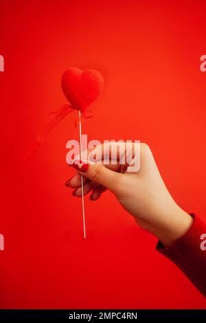 Dekoratives Herz auf einem Stock mit Band in der Hand der Frau. Heller Hintergrund mit silbernen Pailletten und runden Konfetti. Sovereign Samtherzen auf a r Stockfoto