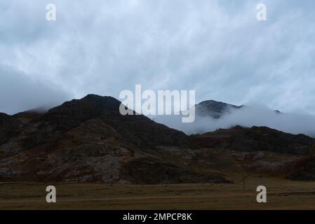 Der Nebel geht am Berghang hinunter und lässt am Abend im Herbst in Altai Schnee auf dem Gipfel nach sich selbst zurück. Blick vom Autofenster. Stockfoto