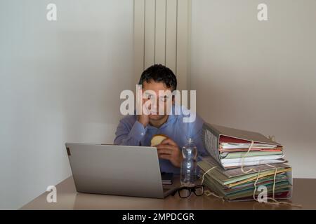 Bild eines verzweifelten Mannes mit einem Sandwich und einer kleinen Flasche Wasser, der wegen Überstunden in seinem Büro arbeiten muss. Stockfoto