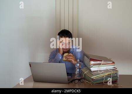 Bild eines verzweifelten Mannes mit einem Sandwich und einer kleinen Flasche Wasser, der wegen Überstunden in seinem Büro arbeiten muss. Stockfoto