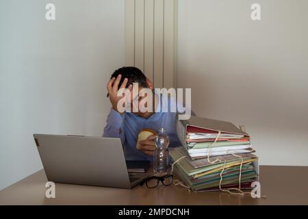 Bild eines gestressten und verzweifelten Geschäftsmanns mit Sandwich und einer Flasche Wasser in der Hand, der für Überstunden im Amt bleiben muss. Stockfoto