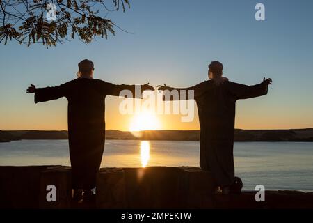 Zwei junge arabische Männer mit offenen Armen, die Djellaba und Turban tragen und auf den Sonnenuntergang vor einem See schauen Stockfoto