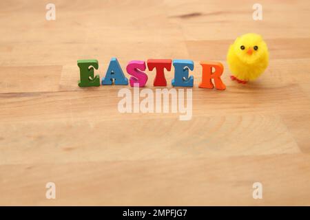 Osterspelz in bunten Holzbuchstaben auf einem natürlichen beigen Holzhintergrund mit einem flauschigen gelben Küken daneben. Stockfoto