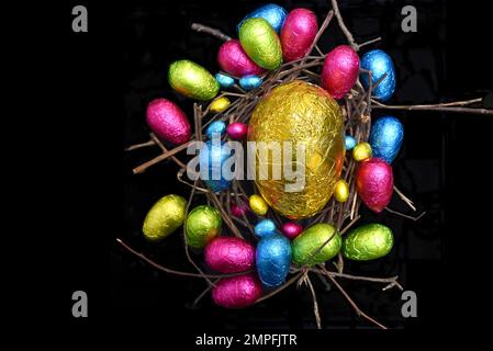In Folie verpackte mehrfarbige ostereier in rosa, grün, blau und gelb in einem natürlichen Nest aus Holz, Stäbchen und Zweigen, vor schwarzem Hintergrund. Stockfoto