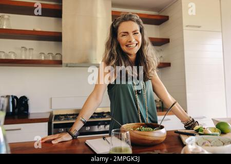 Eine glückliche Veganerin lächelte in die Kamera, während sie eine pflanzenbasierte Mahlzeit zubereitete. Reife Frau, die ein Bio-Rezept in ihrer Küche befolgt. Gesunder Senior wo Stockfoto
