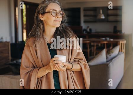 Seniorin blickt mit Bedacht weg, während sie mit einer Tasse Tee in der Hand steht. Reife Frau, die zu Hause eine ruhige Pensionierung genießt. Stockfoto