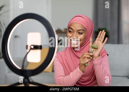 Eine glückliche schwarze, muslimische Bloggerin, die eine Bewertung der Steinrolle macht und ein Video auf einem Smartphone mit Ringlampe filmt Stockfoto