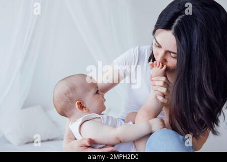 Junge, schöne Mutter küsst ihr süßes Baby auf die Wange. Weiße Brünette berührt ihr lächelndes Kind mit dem Gesicht. Hübsches Baby in einem weißen Body mit H. Stockfoto