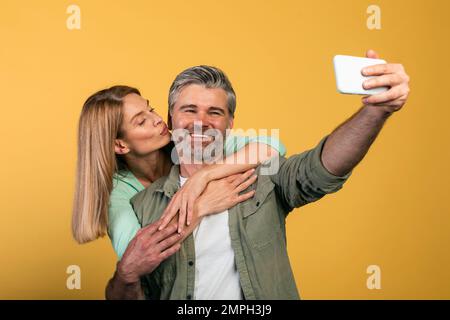 Glückliche Frau mittleren Alters, die ihren Ehemann auf die Wange küsst, Ehepartner, die Selfie auf gelbem Hintergrund machen, Studioaufnahme Stockfoto