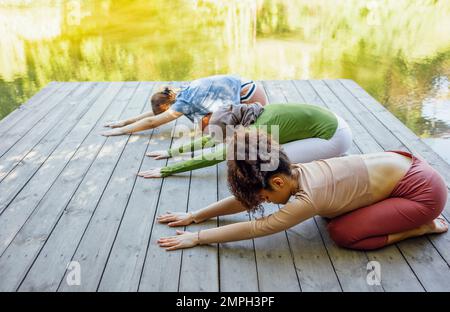 Eine Gruppe von Teenagern trainiert Fitness auf dem Pier am See oder Fluss. Junge und positive Mädchen gehen zum Sport. Moslem in einem Hidschab, afroamerikaner A Stockfoto