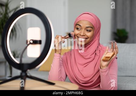 Eine glückliche schwarze moslem-Lady-Beauty-Bloggerin, die Videos für ihre Follower auf einem Smartphone aufnimmt und Make-up aufträgt Stockfoto