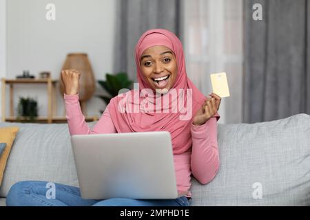 Überglücklich afroamerikanische Frau in Hijab mit Kreditkarte, Einkaufen im Online-Shop auf Laptop, Gesten JA Stockfoto
