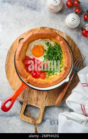 Holländischer Babypfannkuchen. Frischer hausgemachter hausgemachter holländischer Baby-Pfannkuchen mit Spiegelei, Tomate und grünem Rucola in einer gusseisernen Schilfpfanne auf hellgrauem Beton, rustikaler Tabulator Stockfoto