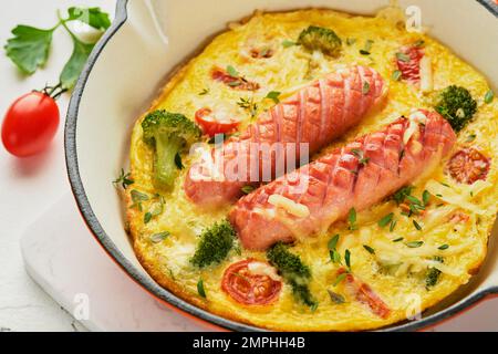 Omelette oder Frittata mit zwei Würstchen, geschmolzenem Käse, Kirschtomaten, Brokkoli und Petersilie auf roter Pfanne oder Eisentopf auf weißem, altem Betonrücken Stockfoto