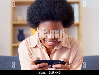 Frau, die zu Hause in der Lounge ein mobiles Spiel auf ihrem Handy im Querformat spielt. Aufgeregter junger Gamer, der lächelt und lacht, während er Spaß hat Stockfoto