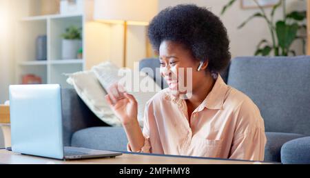 Junge Frau, die mit ihrem Laptop zu Hause sitzt und ihre kabellosen Kopfhörer verwendet. Fröhliche und wunderschöne afroamerikanische Frau mit Afro Stockfoto