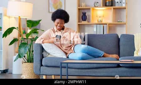 Lachende Frau, die Musik hört und eine Nachricht mit lustigen Memen auf ihrem Handy sendet und Kaffee trinkt, während sie auf dem Sofa sitzt. Glückliche schwarze Frau Stockfoto