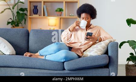Lachende Frau, die Musik hört und eine Nachricht mit lustigen Memen auf ihrem Handy sendet und Kaffee trinkt, während sie auf dem Sofa sitzt. Glückliche schwarze Frau Stockfoto