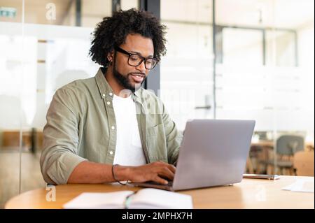 Fokussierter, geschwungener, gutaussehender Mann, der einen Laptop in einem modernen Gemeinschaftsraum verwendet, freiberuflicher Programmierer mit Brillen, der am Schreibtisch sitzt, Kreativitätsdesigner oder Architekt, der schreibt Stockfoto