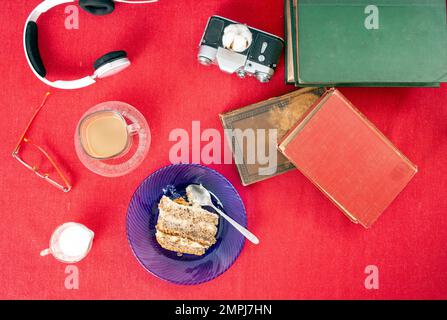 Vintage-Bücher, Kuchenscheiben, Kaffeetassen, Kopfhörer und Gläser liegen flach auf rotem Tischtuch Stockfoto