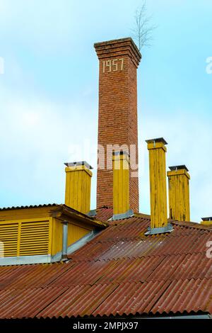 Das Dach eines alten Hauses mit vielen Schornsteinen. Viele kleine Holzschornsteine und ein großer aus Ziegeln mit einer Jahreszahl. Stockfoto