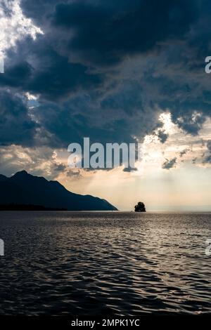 Dunkle Sturmwolken über dem Genfer See, einem einzelnen Baum auf einer kleinen Insel und Berglandschaft in der Ferne. Stockfoto